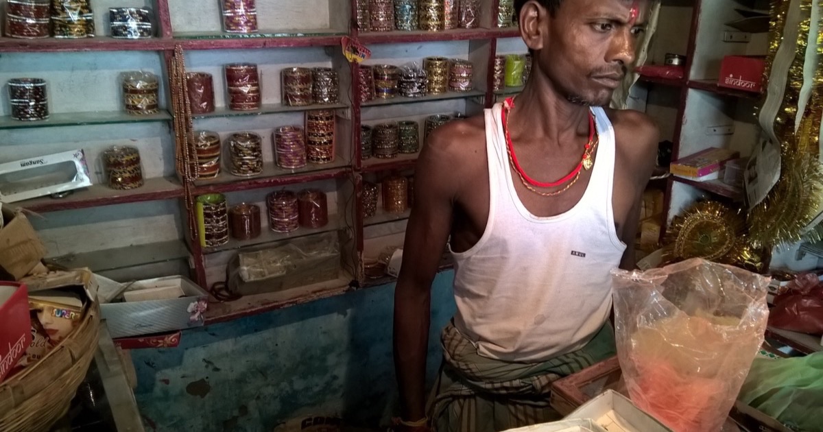 A shopkeeper standing behind the counter of a small Indian shop, shelves of stock behind him